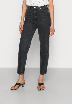 Woman wearing high-waist black denim jeans and black open-toe kitten heel sandals, standing against a plain white background.