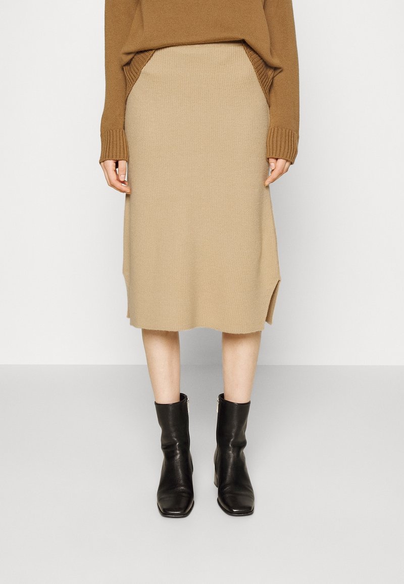 Stickad beige kjol med en figursydd silhuett, medellång längd, sidslitsar och ribbat mönster, matchad med svarta ankelskor.