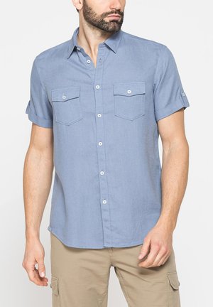 Uomo che indossa una camicia azzurro chiaro a maniche corte con tasche sul petto e pantaloni cargo beige su uno sfondo semplice.