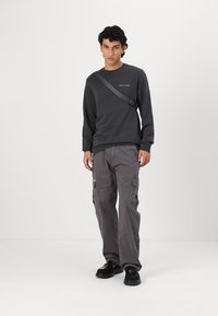 Sudadera gris oscuro con cuello redondo y una sutil marca, combinada con pantalones cargo gris claro que cuentan con bolsillos laterales y un corte relajado. Calzado negro.