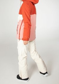 Chaqueta color bloqueada en naranja y rosa con capucha, bolsillos con cremallera y puños elásticos, combinada con pantalones cargo blancos y zapatos negros.