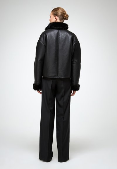 Giacca di pelle nera con colletto e polsini di pelliccia, caratterizzata da un design dritto e accorciato. Abbinata a pantaloni neri a gamba larga.