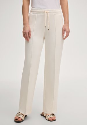 Frau trägt cremefarbene, weit geschnittene, hoch taillierte Hosen mit vorderen Nähten und Kordelzug, kombiniert mit weißen Sandalen mit schwarzem Text.