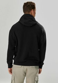Hanorac negru cu o textură moale, având o glugă mare și manșete ribbed. Modelul poartă pantaloni cargo bej cu buzunare vizibile.