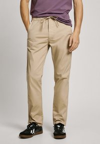 Pantalones de algodón beige con cintura el�ástica y cordón ajustable. Diseño de pierna recta con bolsillos laterales. Combinados con zapatillas negras.