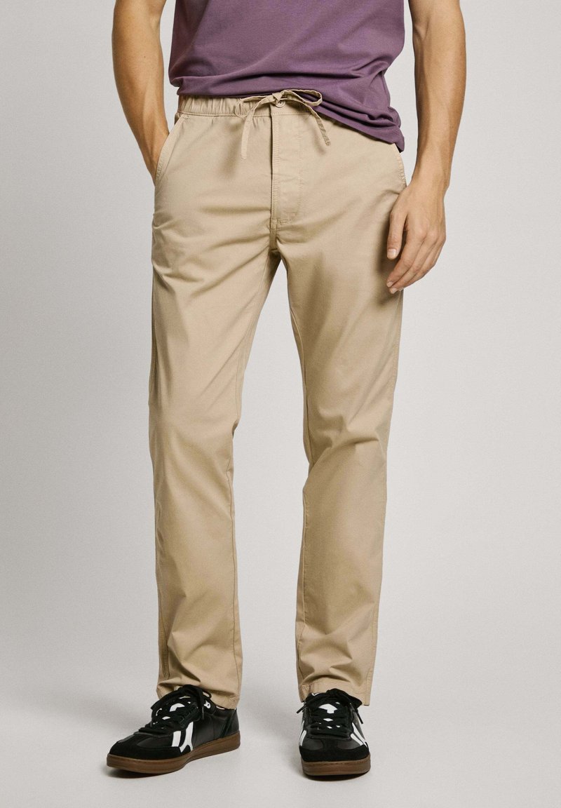 Pantalones de algodón beige con cintura elástica y cordón ajustable. Diseño de pierna recta con bolsillos laterales. Combinados con zapatillas negras.