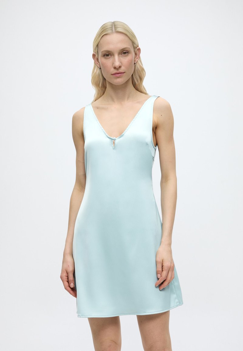 Femme blonde portant une robe en satin bleu clair sans manches avec un col en V et un petit bouton à l’avant, debout devant un fond blanc uni.