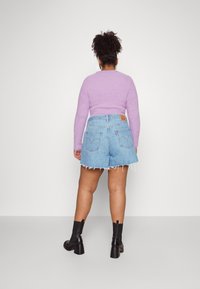 Top de manga larga púrpura y peludo, combinado con pantalones cortos de mezclilla de tiro alto, de color azul claro, con dobladillos deshilachados y una etiqueta de cuero marrón en la parte de atrás. Botas negras de tobillo.