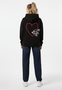 Felpa nera con design a cuore rosso e spille di sicurezza bianche sul retro, abbinata a pantaloni navy con fantasia e sneakers bianche.