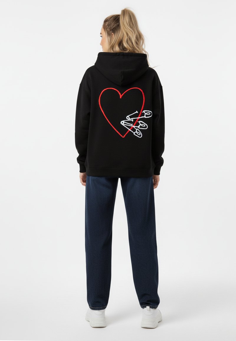 Felpa nera con design a cuore rosso e spille di sicurezza bianche sul retro, abbinata a pantaloni navy con fantasia e sneakers bianche.