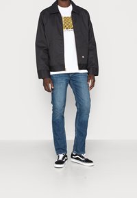 Svart lättviktsjacka med dragkedja, tillsammans med en vit t-shirt med gul text, blå jeans och svartvitt sportskor.