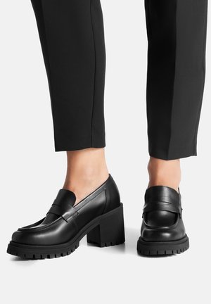 Černé kožené mokasíny se širokým páskem, robustní texturovanou podrážkou a blokovým podpatkem navrženým pro stabilitu.