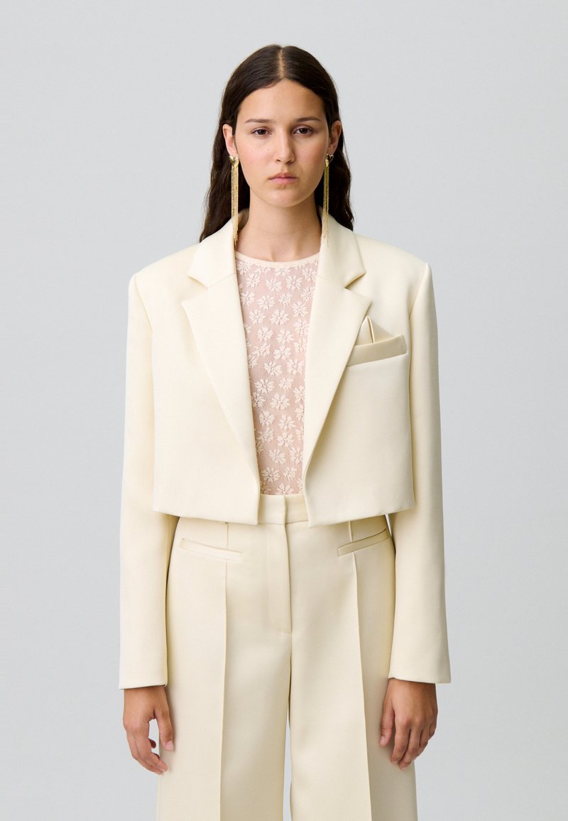 Veste courte crème en tissu structuré, associée à un top en dentelle florale et à un pantalon large plissé de la même couleur.
