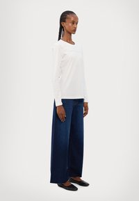 T-shirt bianca a maniche lunghe con scollo rotondo, abbinata a jeans a gamba larga blu scuro. Il modello indossa scarpe basse nere, in piedi su uno sfondo semplice.