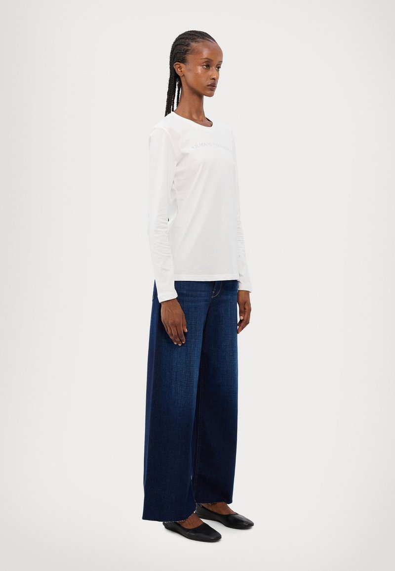 T-shirt bianca a maniche lunghe con scollo rotondo, abbinata a jeans a gamba larga blu scuro. Il modello indossa scarpe basse nere, in piedi su uno sfondo semplice.