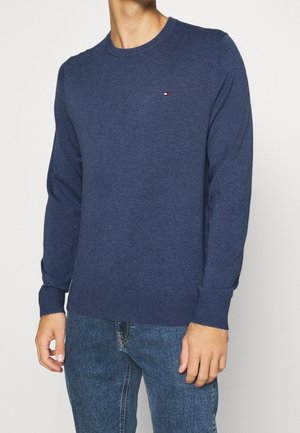 Homme portant un pull bleu marine uni à manches longues avec un petit logo sur la poitrine, associé à un jean en denim bleu.
