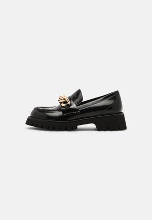 Mocassins en cuir verni noir avec une semelle noire épaisse et un emblème doré sur le devant, présentant une texture lisse et des lignes épurées.
