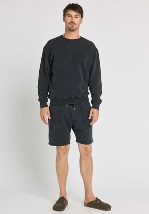 Homme debout portant un sweat-shirt noir à manches longues, un short noir avec des poches avant et des chaussures sombres sans lacets sur fond uni.