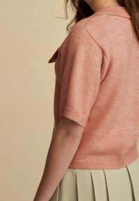 Sweter z krótkim rękawem w miękkim koralowym splocie, z złożonym kołnierzykiem, prążkowanym wykończeniem i małym guzikiem na ramieniu.