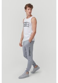 White tank top with black logo design, paired with grey sweatpants featuring a side logo. Grey sneakers complete the look.