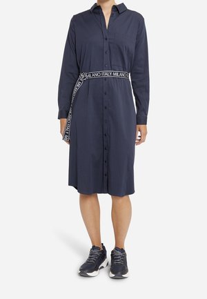 Abito camicia lungo blu navy con maniche lunghe, bottoni frontali e tasca sul petto, cinte in vita con una cintura a righe bianche e nere.