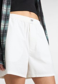 Karierte weiße Shorts mit einem elastischen Bund, die über eine dezente Textur und ein kleines Label-Logo am hinteren Bund verfügen, kombiniert mit einem karierten Hemd.
