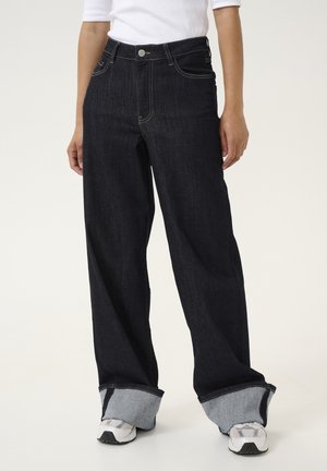 Personne portant un jean en denim foncé à jambes larges avec de grands revers et des baskets blanches, debout devant un fond uni.