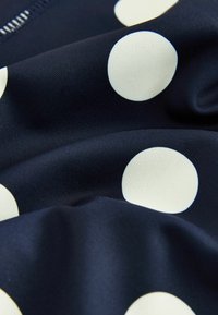 Tissu bleu marine avec de grands pois blancs et une texture visible, montrant de légers plis et un détail partiel de fermeture éclair.