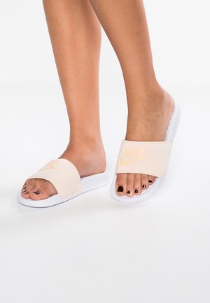 Person iført hvide Nike slide-sandaler med sort neglelak på tæerne, stående på en hvid overflade.