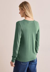 Grüner Langarmpullover mit rundem Halsauschnitt und gerippten Bündchen; weiche Textur; getragen mit blauen Jeans; minimalistisches Design, keine Muster.