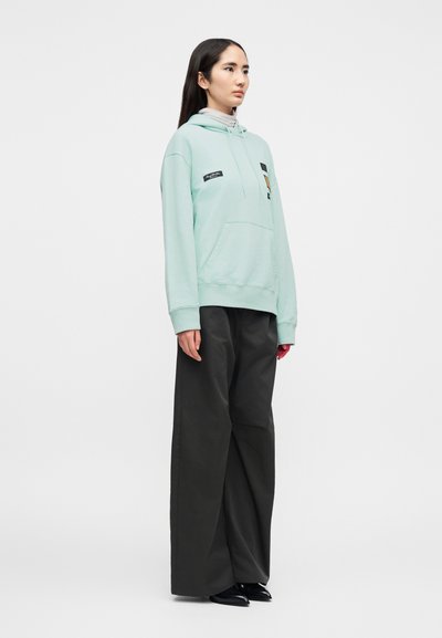 Σ mint πρ άσινο φούτερ με κουκούλα, εμπρός τσέπη και patches, σε συνδυασμό με μαύρο παντελόνι φαρδιάς γραμμής και μαύρα μυτερά παπούτσια, φορώντας το ένα άτομο.