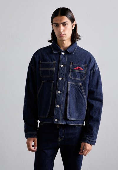 Veste en denim bleu foncé avec un col pointu, fermeture à boutons sur le devant, plusieurs poches et accent logo rouge. Tissu texturé avec des coutures jaunes.