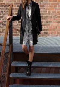 Cappotto nero su un vestito bianco a fantasia con dettagli scuri, abbinato a stivali neri lucidi alla caviglia. La modella scende delle scale di metallo.