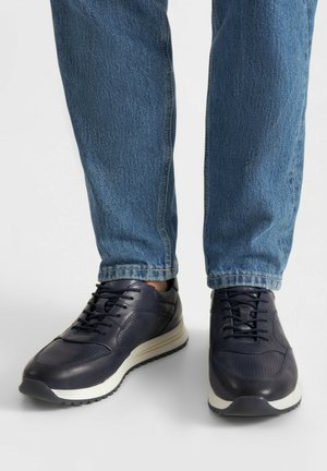 Muž nosící modré kožené tenisky s bílými podrážkami a modré džínové kalhoty, stojící na bílé ploše.