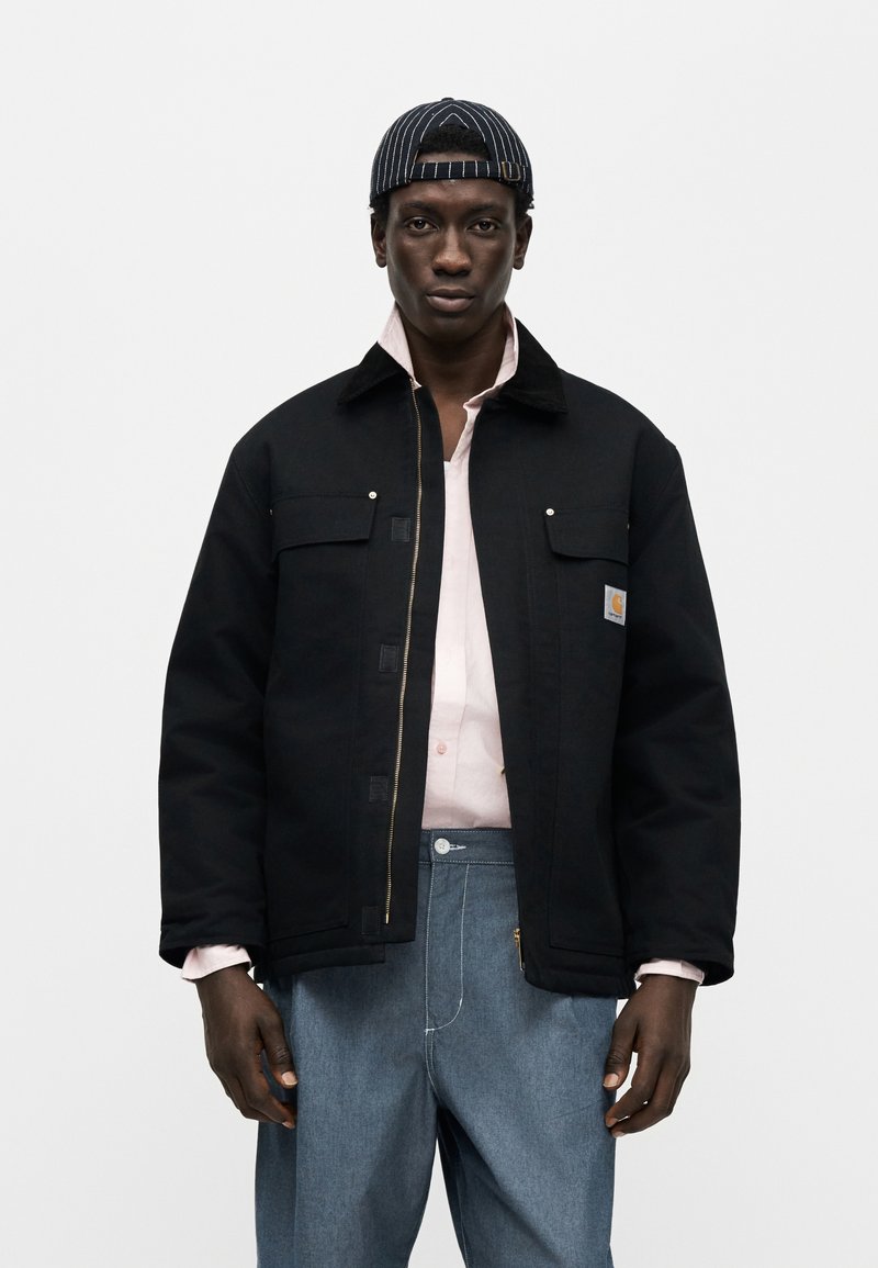 Zwarte katoenen jas met een ritssluiting aan de voorkant, borstzakken en metalen hardware. Draagt over een lichtroze shirt en gecombineerd met blauwe spijkerbroek.