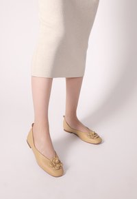 Beige Lederflats mit einer abgerundeten Zehenpartie, die an der Vorderseite mit einem floralen Akzent verziert sind und einen niedrigen, braunen Gummihügel haben. Eine gerippte cremefarbene Rock ist sichtbar.