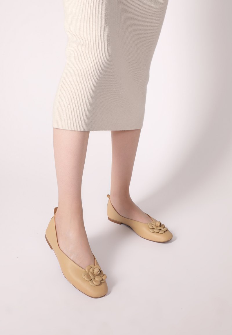 Beige Lederflats mit einer abgerundeten Zehenpartie, die an der Vorderseite mit einem floralen Akzent verziert sind und einen niedrigen, braunen Gummihügel haben. Eine gerippte cremefarbene Rock ist sichtbar.