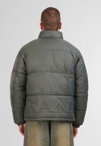 Tumedatextuuriline soe jakk tumehalli värviga, millel on kõrge krae, quiltitud osad ja reljeefne "KAN" logo seljal. Sujuv tekstuur.