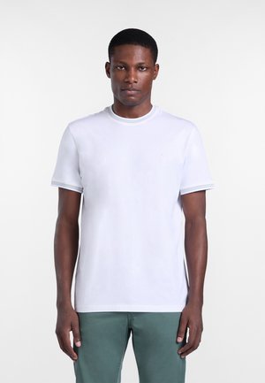 Muž stojaci čelom vpred, oblečený v jednoduchom bielom tričku s krátkym rukávom a zelených nohaviciach, na bielom pozadí.