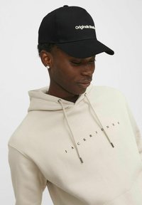 Mustmust värvi kork, millel on valge brodering "Originals Studio"; beež kapuutsiga pusa, millel on must trükk "JACK & JONES"; sile kangas, mugav lõige.