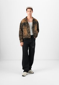 Chaqueta de denim marrón con textura desgastada, pantalones vaqueros de pierna ancha negros con detalles de tachuelas y zapatillas neutras. Conjunto simple y casual.