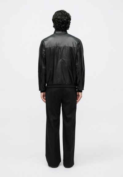 Crna kožna jakna glatkog završetka s rebrastim manšetama, uparena s širokim crnim hlačama. Model je prikazan straga.