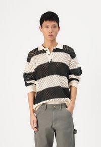 Pull en tricot avec motif en maille ouverte, featuring des rayures horizontales noires et crème, un col et des manches retroussées ; associé à un pantalon gris.