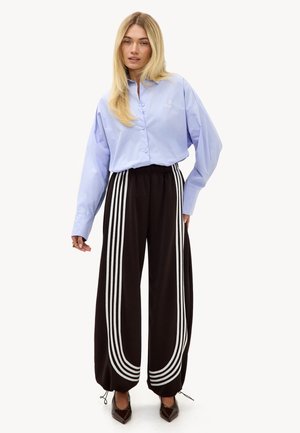 Femme portant une chemise bleue claire boutonnée et un pantalon noir à jambes larges avec des rayures blanches, debout avec une main légèrement levée.