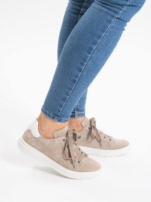 Person trägt hellbraune Wildleder-Sneaker mit weißen Sohlen und grauen Schnürsenkeln, kombiniert mit blauen Skinny-Jeans aus Denim vor weißem Hintergrund.