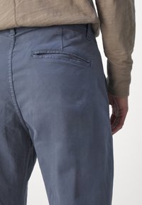 Gros plan d'une personne portant un pantalon bleu avec une seule poche passepoilée à l'arrière, associée à une chemise beige à manches longues.