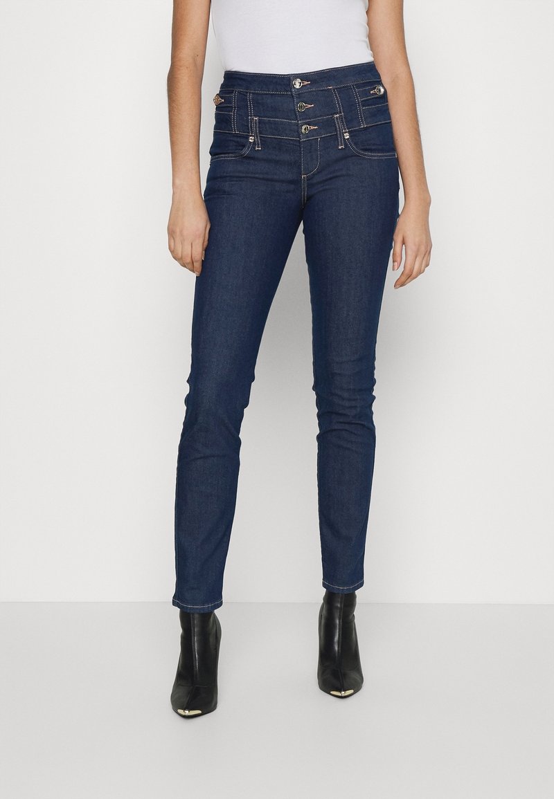 Jeans ajustados de mezclilla azul oscuro, con una cintura alta, cierre de varios botones y costuras en contraste. Combinados con botas negras hasta el tobillo.