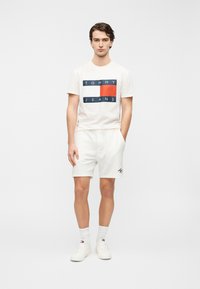 Άνδρικό λευκό βαμβακερό t-shirt με λογότυπο "TOMMY JEANS" σε ναυτικό μπλε και κόκκινο, συνδυασμένο με λευκά βαμβακερά σορτς, λευκές κάλτσες και λευκά αθλητικά παπούτσια.