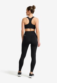 Kvinna som står vänd bakåt iklädd svart sport-bh, högmidjade svarta leggings och svarta löparskor mot en vit bakgrund.