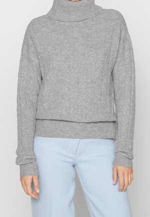 Vrouw draagt een lichtgrijze kabelgebreide coltrui met geribbelde manchetten en zoom, gecombineerd met lichtblauwe broek.